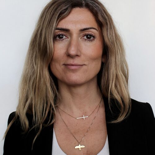 Aralia Eriksson, long brown hair, olive skin wearing black suit and white shirt with two gold chains