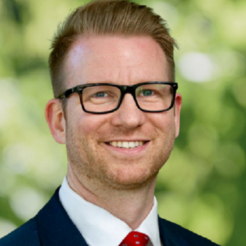 Ayden Sims wearing black glasses, manicured beard, dark suit, white shirt and red tie in front of greenery.