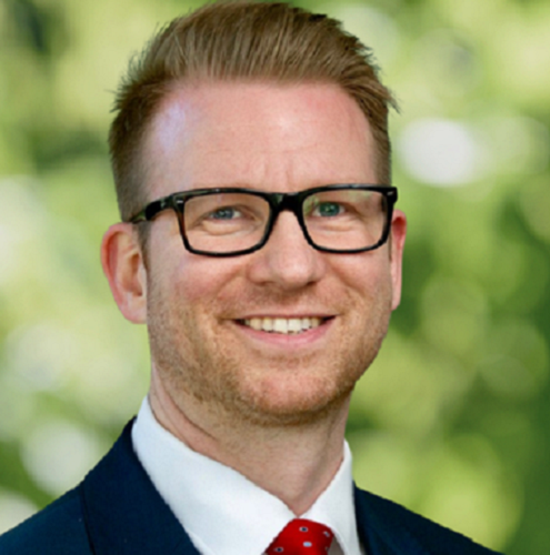 Ayden Sims wearing black glasses, manicured beard, dark suit, white shirt and red tie in front of greenery.