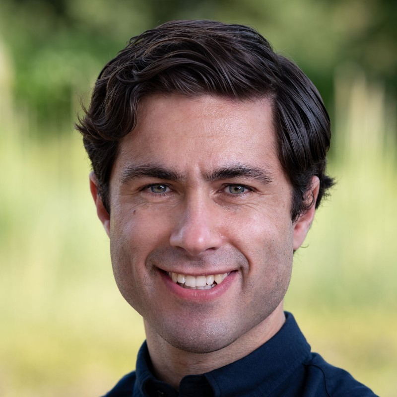 Oli Goodarzi smiling at camera wearing blue shirt and with greenery in the background.