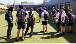 Group of young people in school uniform with backpacks listen to two people from Purple Hands.