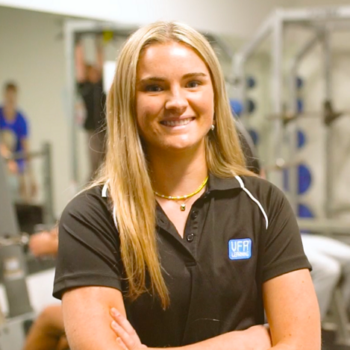 Hannah with long blonde hair, wearing a brown gym shirt smiles into camera with crossed arms.