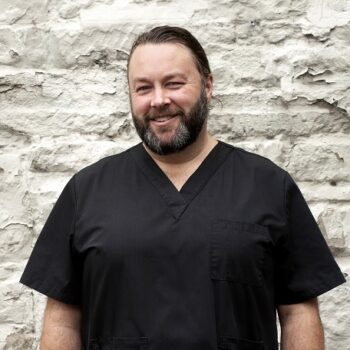 Tim, with dark hair and a beard, wears black nurses' scrubs in front of a white brick wall.