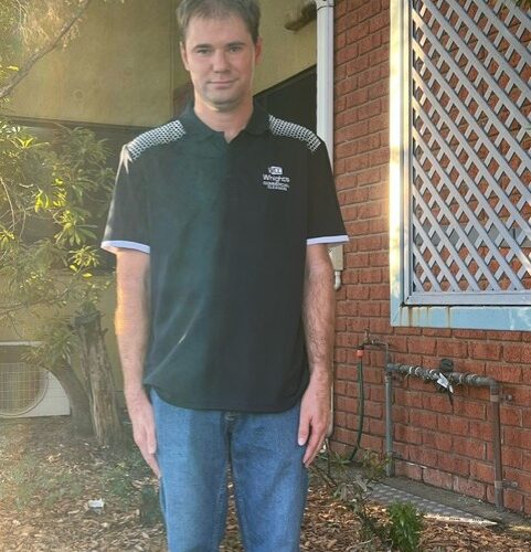Young man with dark hair, dressed in a dark shirt and jeans, stands in front of a brick building.